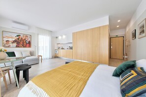 A modern hotel room with a bed, a sofa, a small dining table, and a kitchenette.
