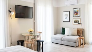 A modern hotel room with a flat-screen TV, a round glass table with chairs, a sofa, and a small side table with books. - Art Home Apartments (Aveiro)