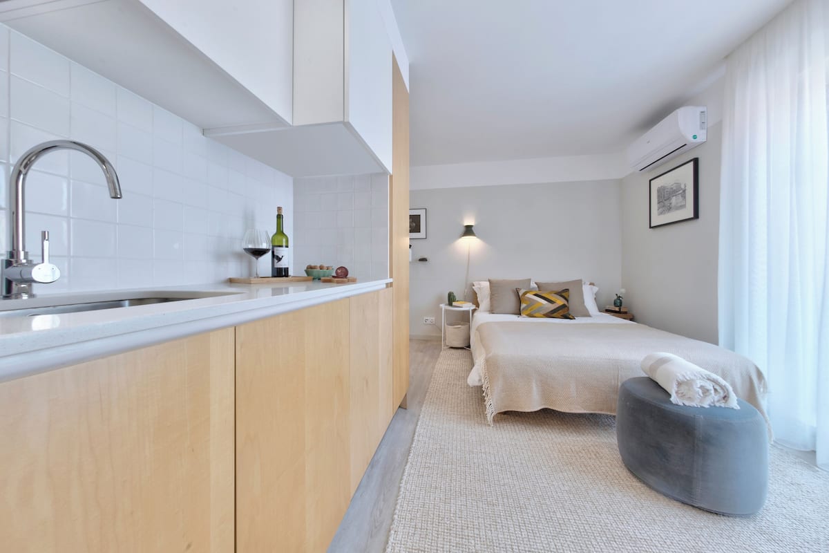 A modern kitchen with a sink, wine bottle, and fruit bowl. A bed with pillows and a blanket is visible in the background.