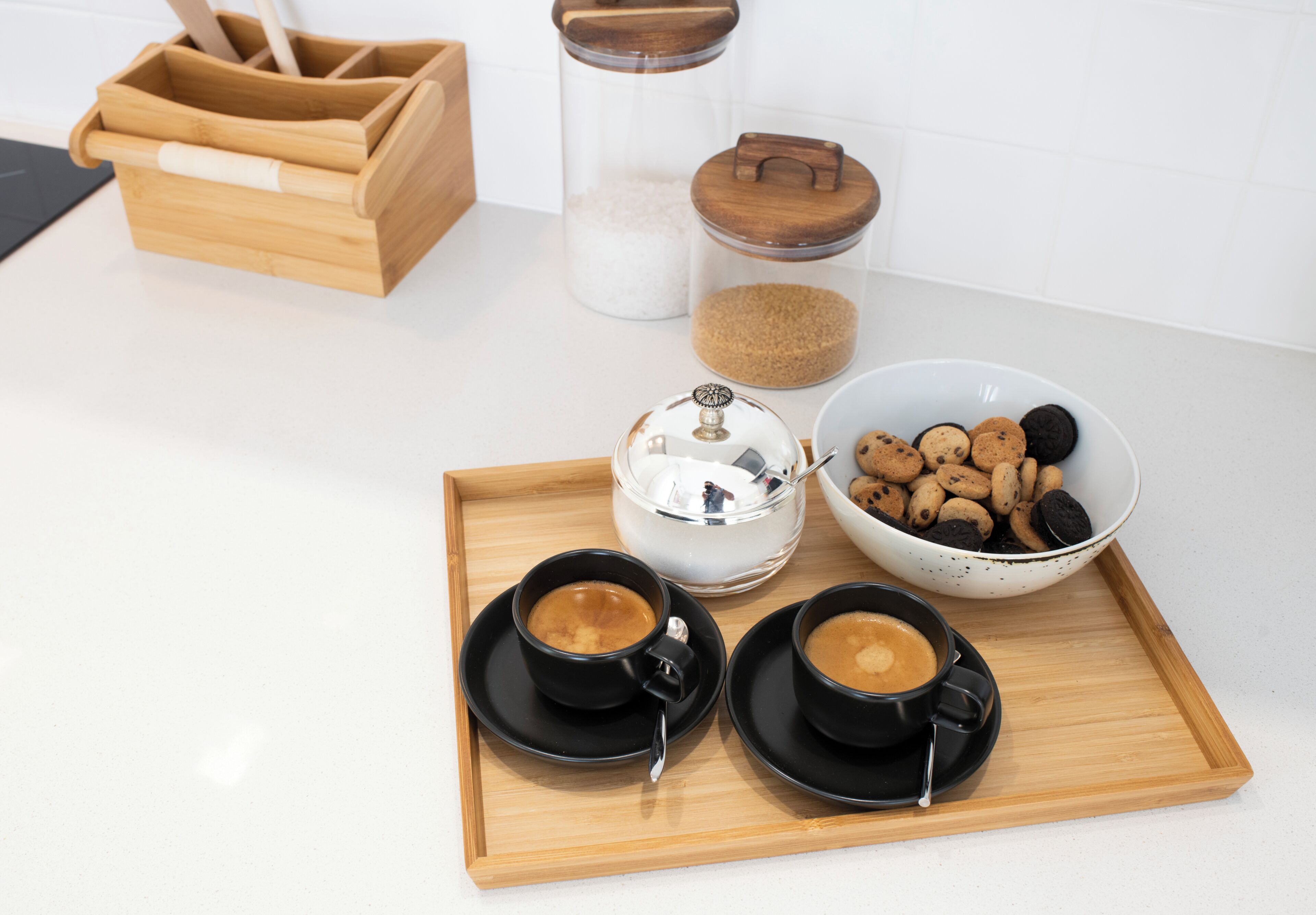 A tray with two cups of coffee, a glass of milk, a bowl of cookies, and two containers of sugar and flour on a kitchen counter.