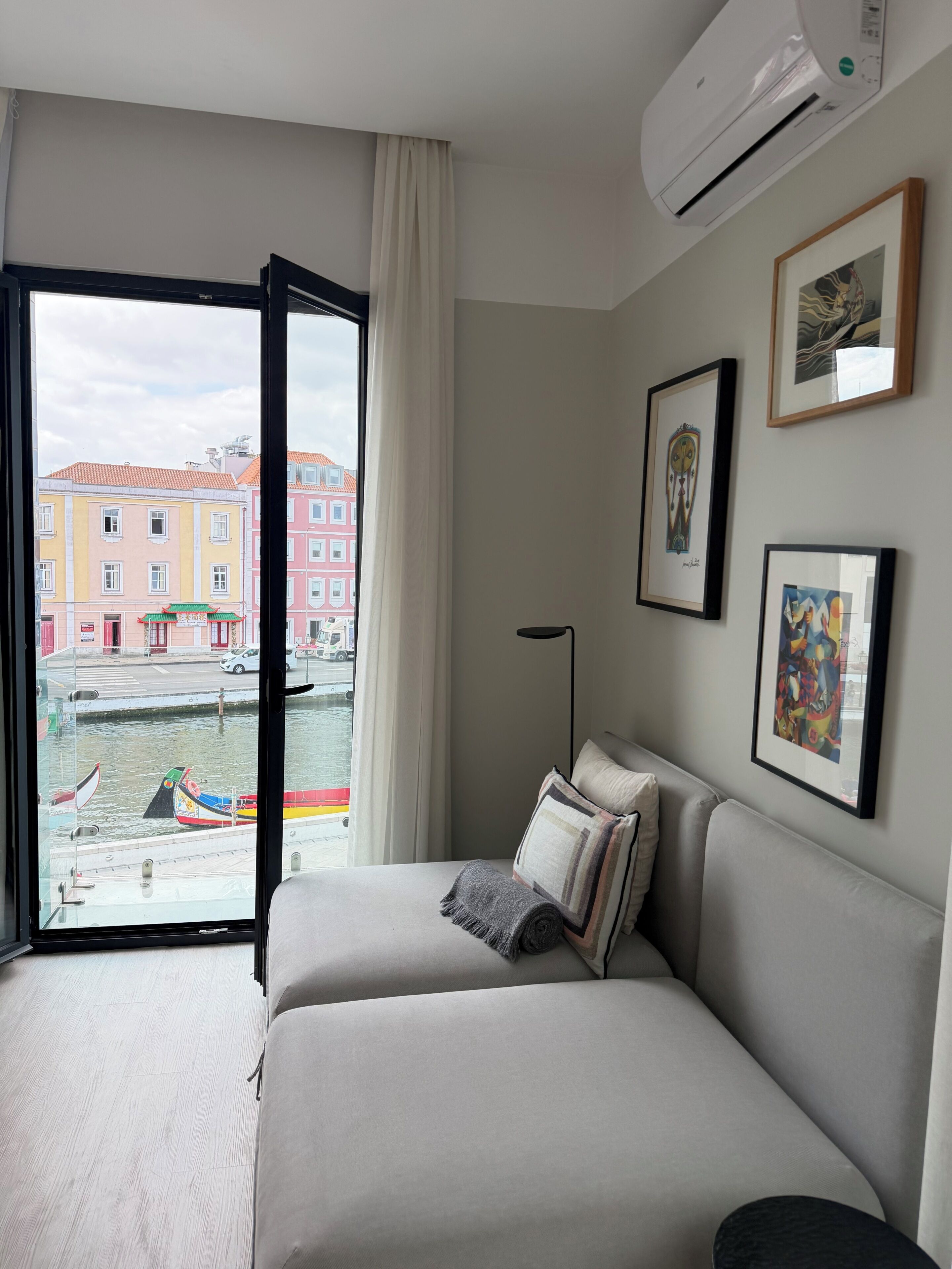 A hotel room with a bed, a window with a view of buildings and boats, and an air conditioning unit.