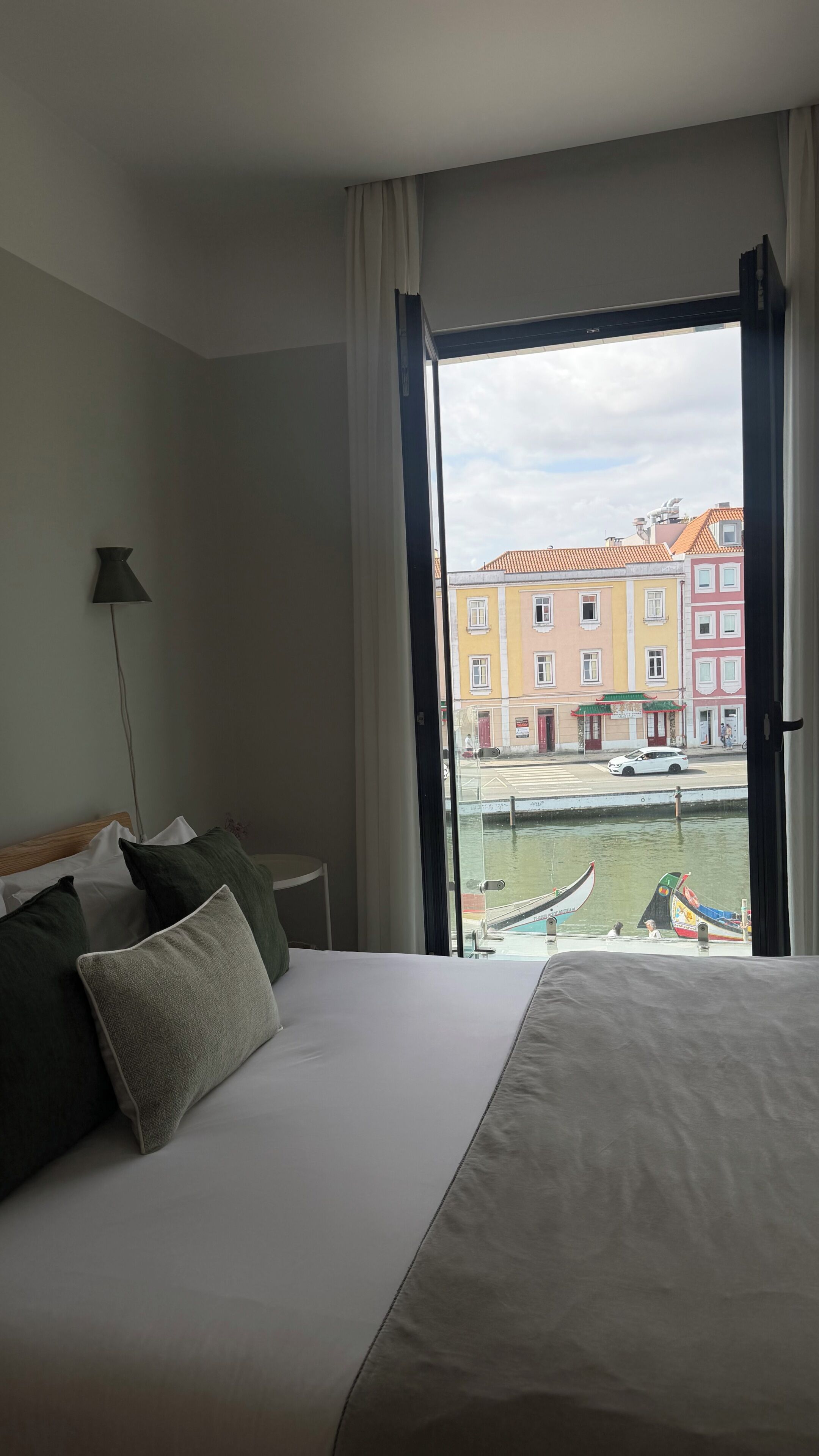 A hotel room with a bed, a nightstand, a lamp, and a view of a street with buildings and a boat.