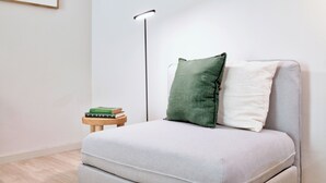 A minimalist room with a grey sofa, a green and white pillow, a small wooden side table with books, and a floor lamp. - Art Home Apartments (Aveiro)