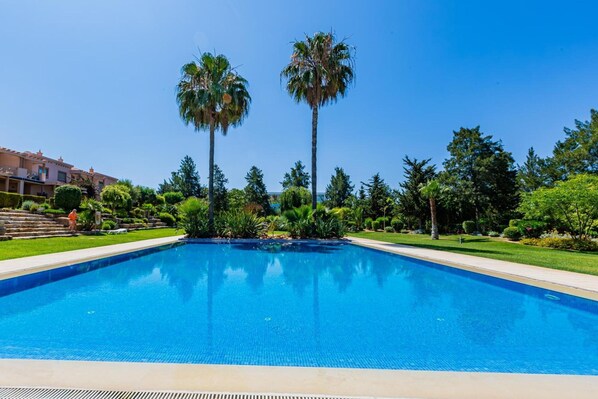 A rectangular swimming pool with clear blue water, surrounded by well-maintained greenery and palm trees. - Casa Colina – Spacious Villa w/ Pool Near Beach! (Albufeira)