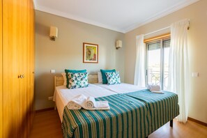 A bedroom with a bed, pillows, a window with curtains, a painting on the wall, and a wooden wardrobe. - Casa Colina – Spacious Villa w/ Pool Near Beach! (Albufeira)