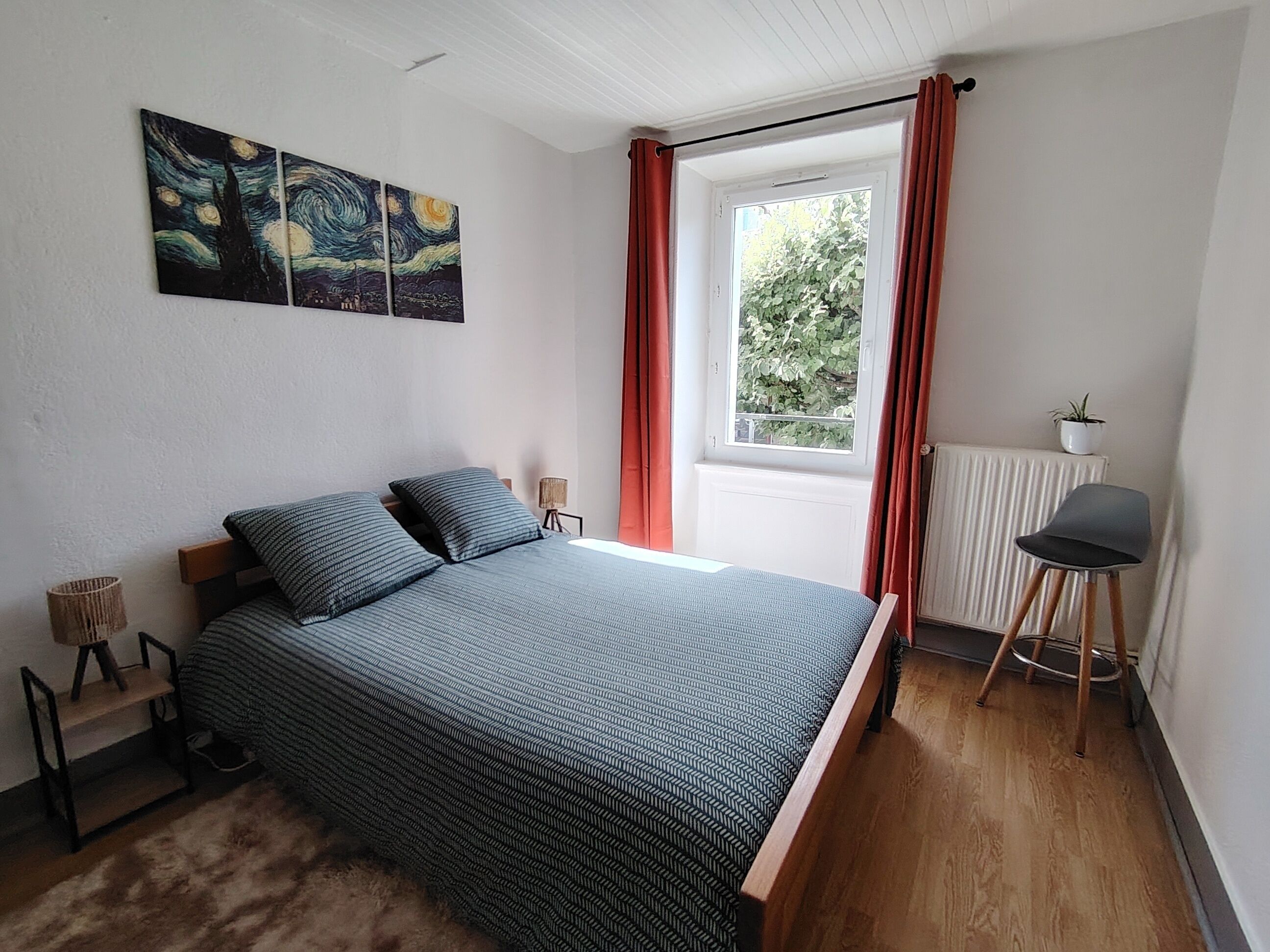 Ein Schlafzimmer mit Bett, einem Fenster mit Vorhängen, einem Heizkörper, einem Stuhl und einem Bild an der Wand.
