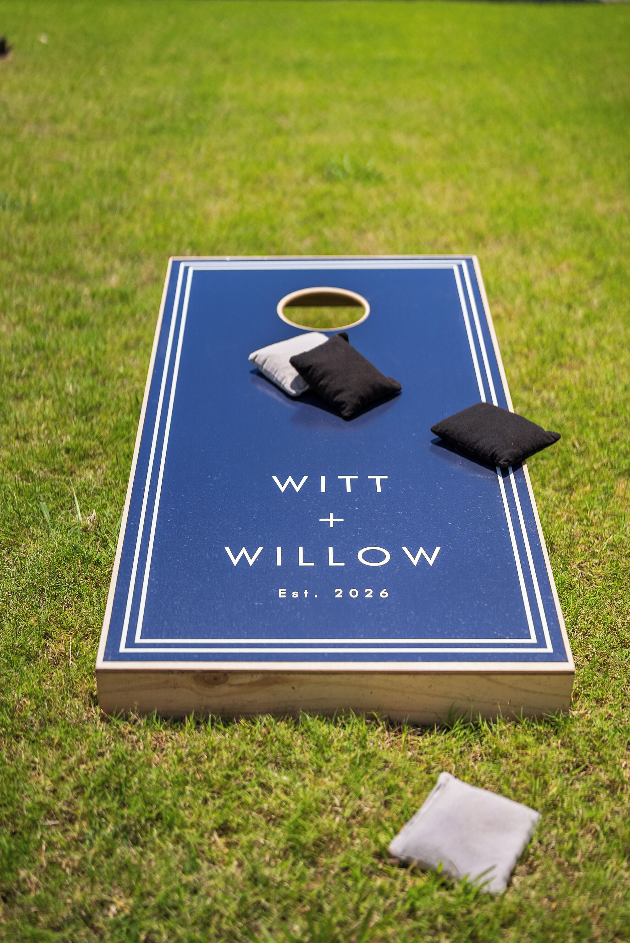 A blue board game with 'WITT + WILLOW' and 'Est 2026' on it, placed on grass.
