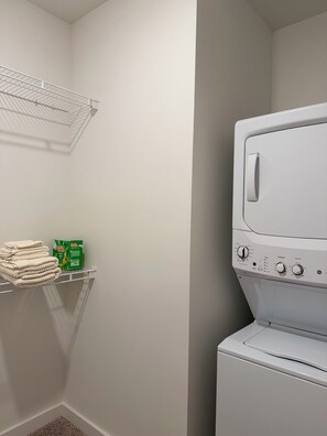 Ein Waschraum mit einer gestapelten Waschmaschine und einem Trockner, einem Regal mit gefalteten Handtüchern und einer Box mit Waschmittel.