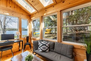 A cabin interior with a wooden interior, a sofa, a desk with a chair, and large windows offering views of the outdoors. - Haliburton Lakefront Escape with Hot Tub, Fire Pit, Sauna & BBQ (Algonquin Highlands)