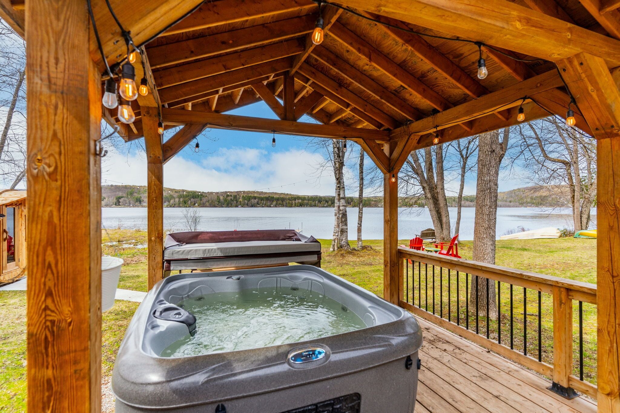 Eine Holzterrasse mit Whirlpool, eine Holzw pergola mit Lichterketten und Blick auf einen See.