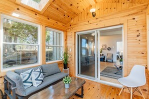 A wooden cabin interior with a sofa, a white chair, a dining table, and a large sliding glass door leading to a dining area. - Haliburton Lakefront Escape with Hot Tub, Fire Pit, Sauna & BBQ (Algonquin Highlands)