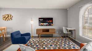 Un soggiorno moderno con una poltrona blu, un mobile TV in legno, un tappeto decorato e una TV a muro che mostra una serie Netflix.