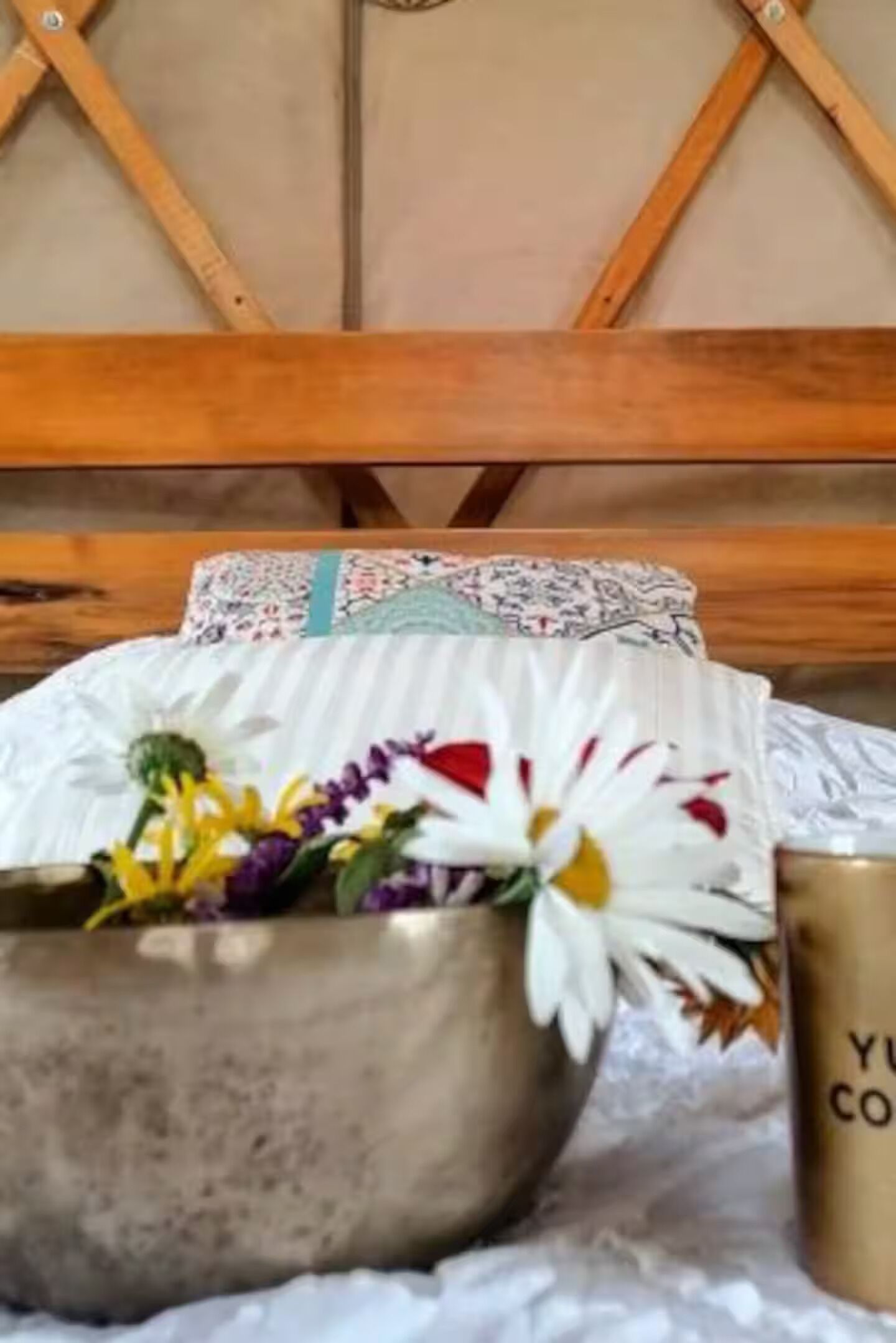 A bed with a wooden headboard, a metal bowl with flowers, and a gold mug on a white cloth.