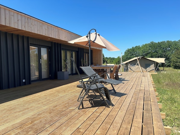 A modern outdoor deck with a dining area, a large umbrella, and a tented structure in the background.