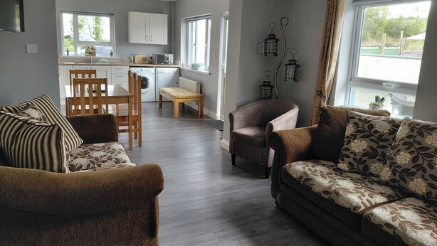 A living room with a sofa, a chair, a dining table, and a kitchen in the background. - Cottage - Sleeps 6 - Parking (Rathmullan)