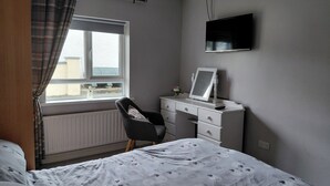 A bedroom with a bed, a desk, a chair, a radiator, a window with curtains, and a TV mounted on the wall. - Cottage - Sleeps 6 - Parking (Rathmullan)