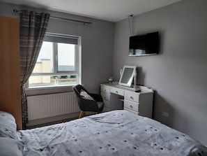 A bedroom with a bed, a desk, a chair, a radiator, a window with curtains, and a TV mounted on the wall. - Cottage - Sleeps 6 - Parking (Rathmullan)