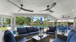 Eine Terrasse im Freien mit Korbmöbeln, Deckenventilatoren und Blick auf einen Pool und Grünflächen.