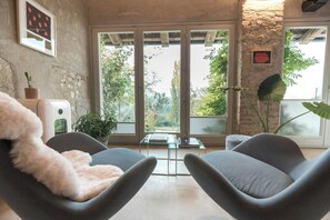 A room with two grey armchairs, a small table, a potted plant, and a view of trees through large windows. - Double-house with pool among Barolo vineyards (Novello)