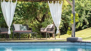 Een zwembadterras met twee stoelen, een tafel en een pergola met witte gordijnen.