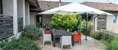 Une terrasse avec une table et des chaises, entourée de gravier, de plantes et d’un parasol blanc.