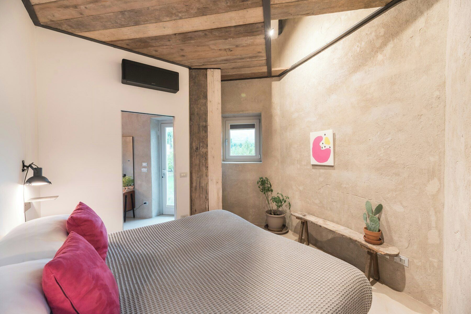 A bedroom with a bed, a potted plant, a small window, and a wall-mounted air conditioner.