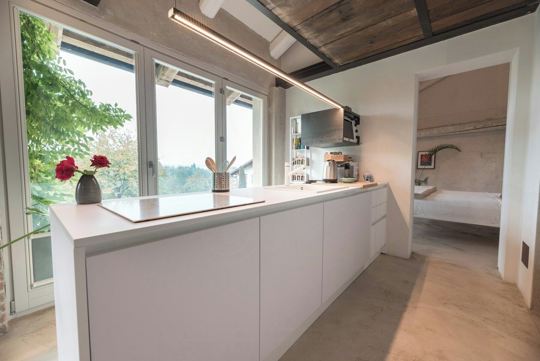 Una cocina moderna con una isla grande, un microondas y un lavamanos. Hay una ventana con vistas a árboles y una cama en la habitación contigua.