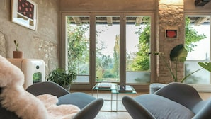 Ein Zimmer mit Steinwand, einem großen Fenster, zwei grauen Stühlen, einem Glastisch und einer Pflanze.