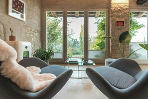 Una habitación con pared de piedra, un ventanal grande, dos sillas grises, una mesa de vidrio y una planta.