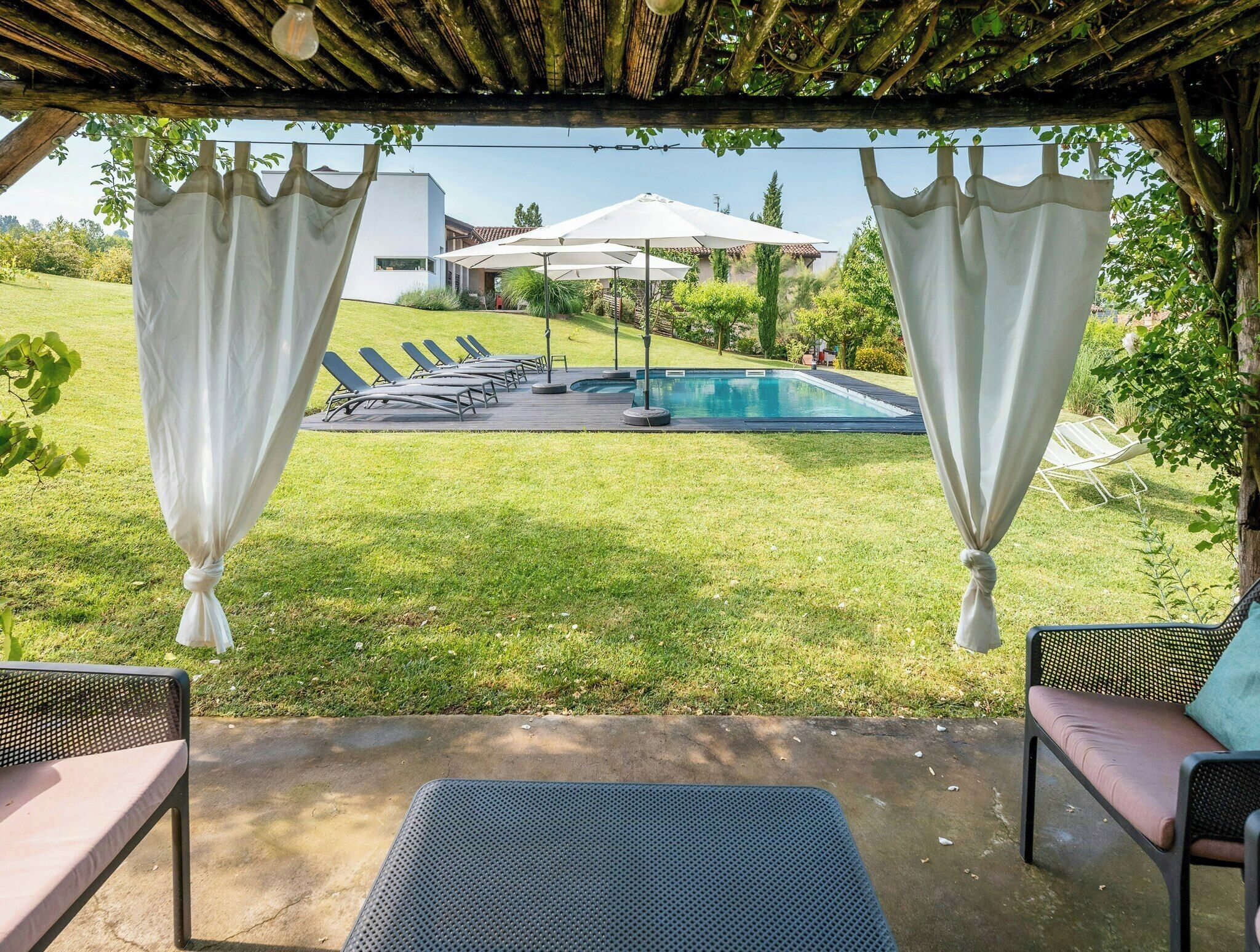 A covered outdoor seating area with a view of a swimming pool, lounge chairs, and a pergola.