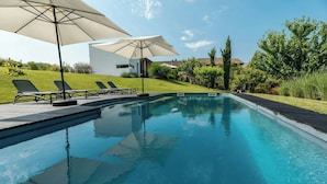 Ein Swimmingpool mit Liegestühlen und Sonnenschirmen, umgeben von viel Grün und einem modernen Gebäude.