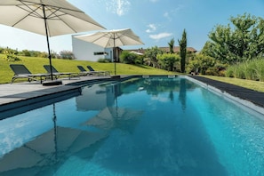 Piscina con sillas de descanso y sombrillas, rodeada de vegetación y un edificio moderno.