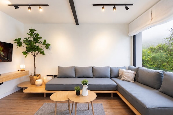 Ein modernes Wohnzimmer mit einem grauen Ecksofa, einem kleinen Holztisch, einem Blumentopf und einem großen Fenster mit Blick auf Grünflächen.