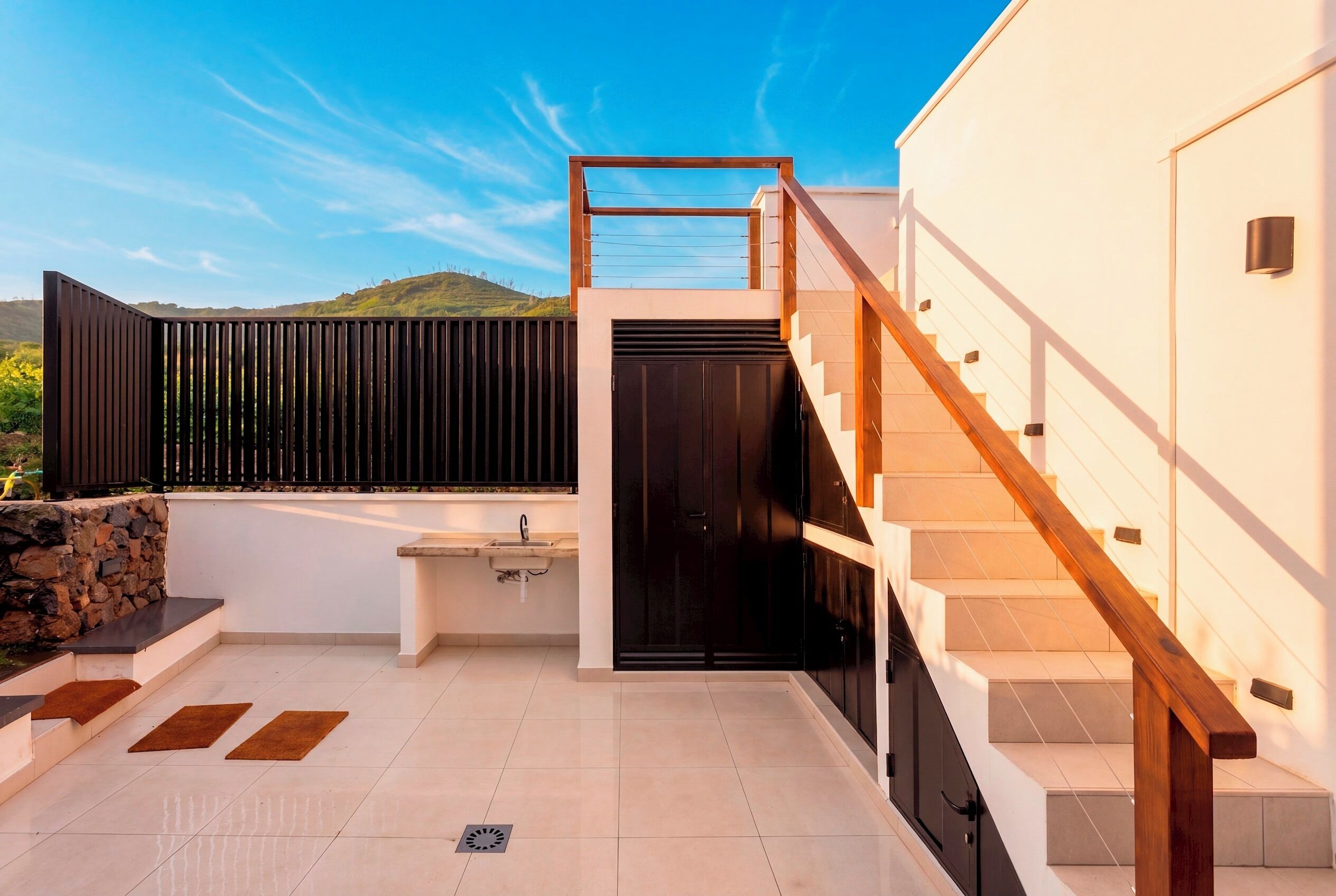 A modern outdoor kitchen area with a sink, a black door, and a staircase leading to an upper level.