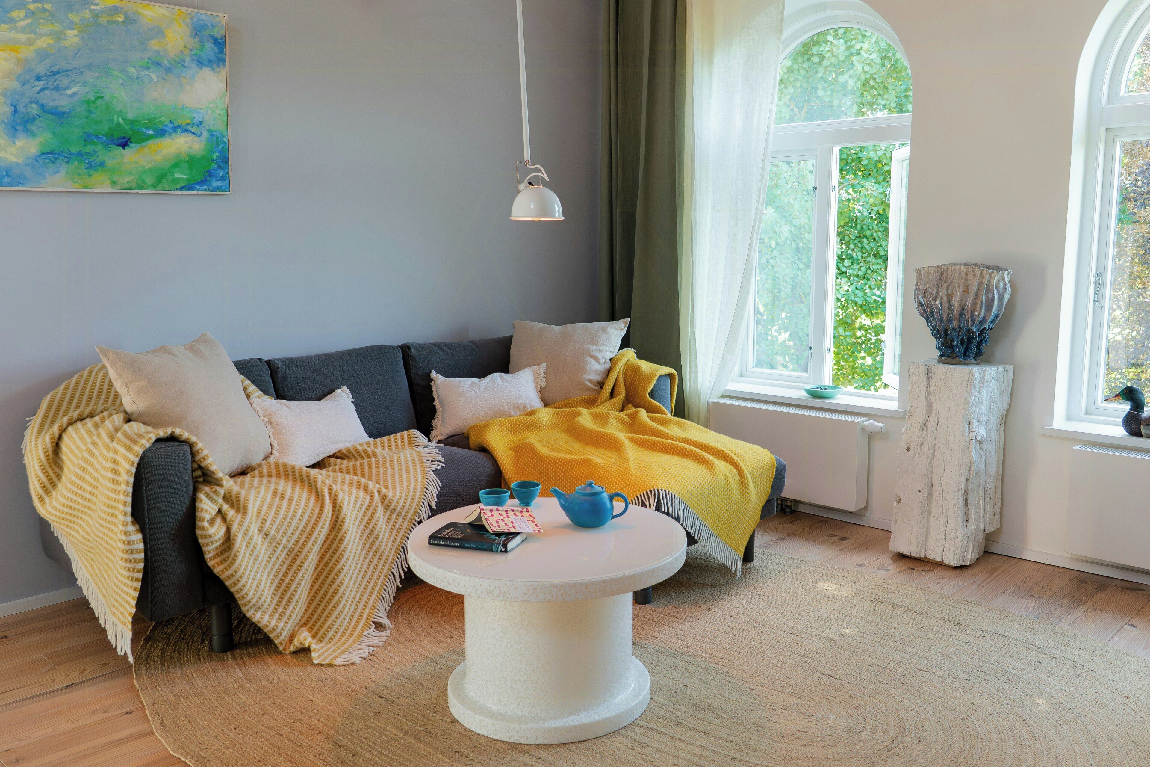 A living room with a sofa, a round coffee table, a painting on the wall, and a window with a view of greenery.