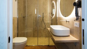 Un baño moderno con ducha de vidrio, lavamanos blanco y un secador de pelo portátil fijado en la pared.