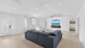 En moderne stue med en stor grå hjørnesofa, et hvidt sofabord, et fladskærms-TV monteret på væggen og et stort vindue med gardiner.