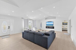 Ein modernes Wohnzimmer mit einem großen, grauen Ecksofa, einem weißen Couchtisch, einem flachbildfernseher an der Wand und einem großen Fenster mit Vorhängen.