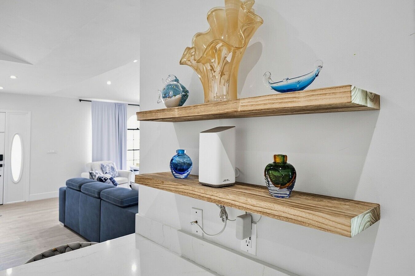 Ein modernes Wohnzimmer mit einem weißen Sofa, einem blauen Pouf und einem Holzregal mit dekorativen Glasvasen.