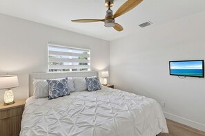 Ein Schlafzimmer mit Bett, Deckenventilator, Fenster mit Jalousien, Fernseher und Nachttischlampen.