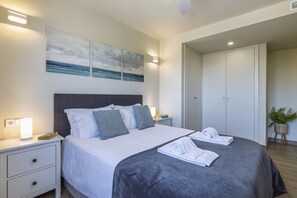 Ein ordentlich bezogenes Bett mit grauem Kopfteil, weißer Bettwäsche und einer grauen Decke. Zwei Nachttische mit Lampen, ein Wandbild und ein Blumentopf sind vorhanden.