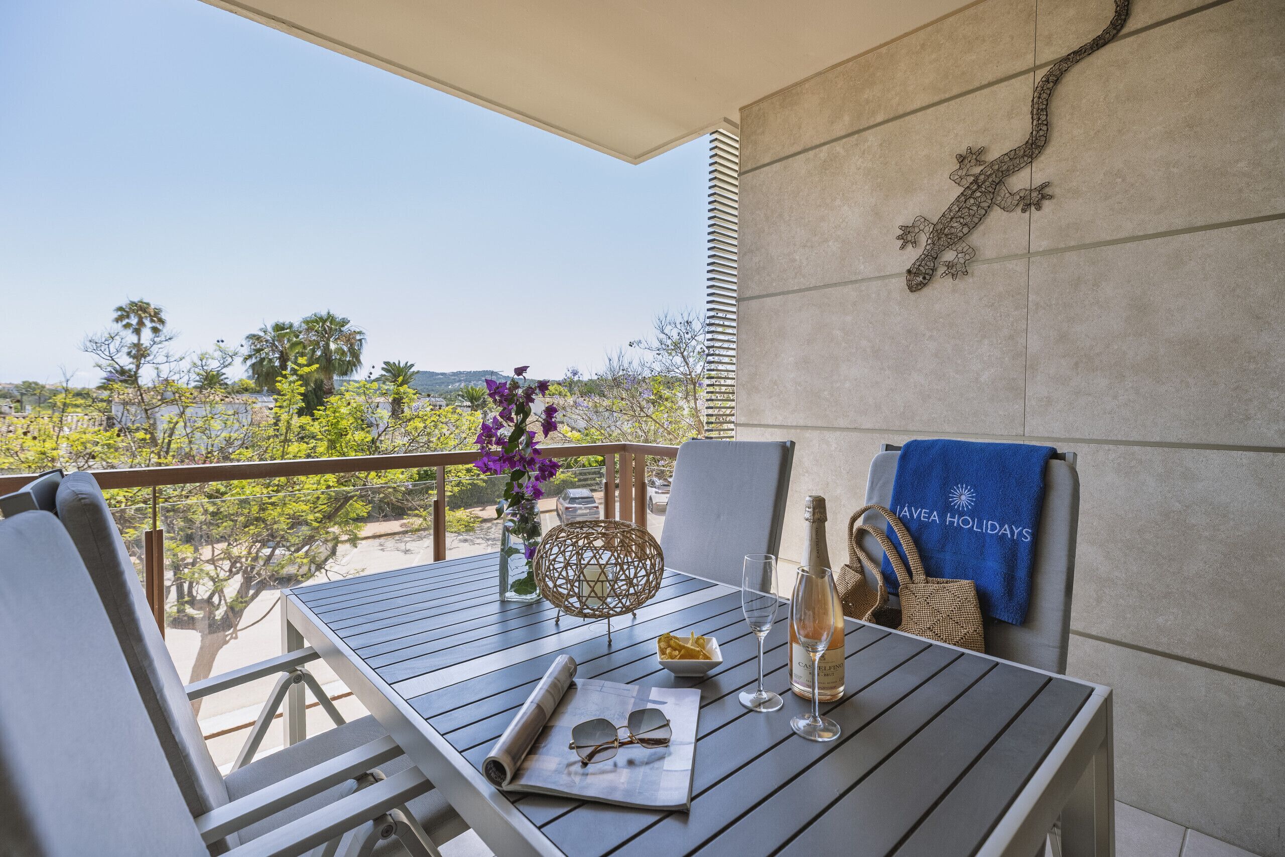 A balcony with a table set for two, a bottle of champagne, a bowl of snacks, and a decorative wall piece.