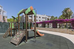 Ein Spielplatz mit Rutsche und Klettergerüsten, umgeben von einer Steinmauer und violetten Blumen.