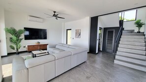 Ein modernes Wohnzimmer mit einem Ecksofa, einem Flachbildfernseher, einem Deckenventilator und einer Treppe.