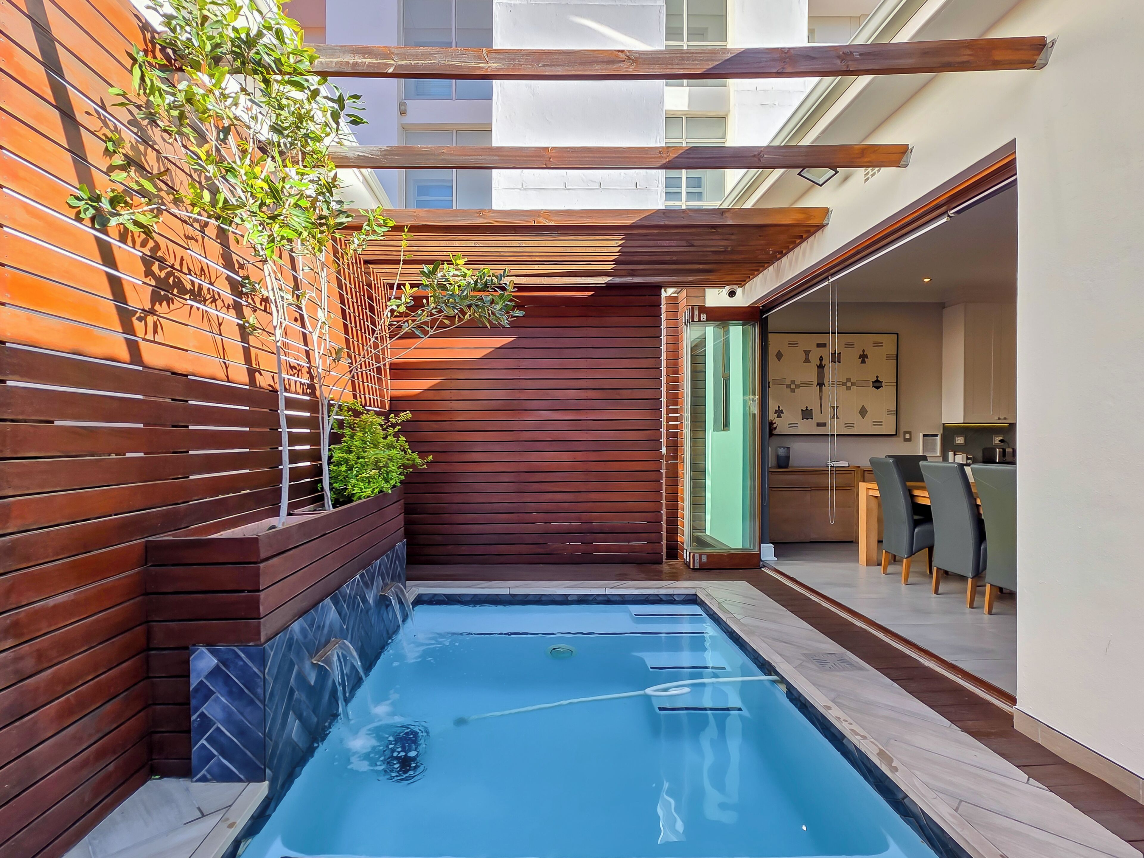Un área de piscina moderna al aire libre con cubierta de madera, puerta de vidrio que da a un comedor y borde de piscina con baldosas.