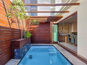 Un área de piscina moderna al aire libre con cubierta de madera, puerta de vidrio que da a un comedor y borde de piscina con baldosas.