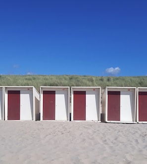 Una hilera de cabañas de playa numeradas con puertas blancas y rojas, sobre arena, bajo un cielo azul despejado.