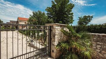 A gated entrance with a stone wall, a palm tree, and a building with a red roof in the background.