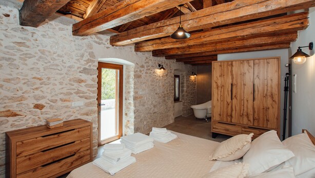 Ein Schlafzimmer mit Steinsäulen, Holzbalken, einem Bett mit weißen Leinen, einer Holzkommode und einem Holzschrank.