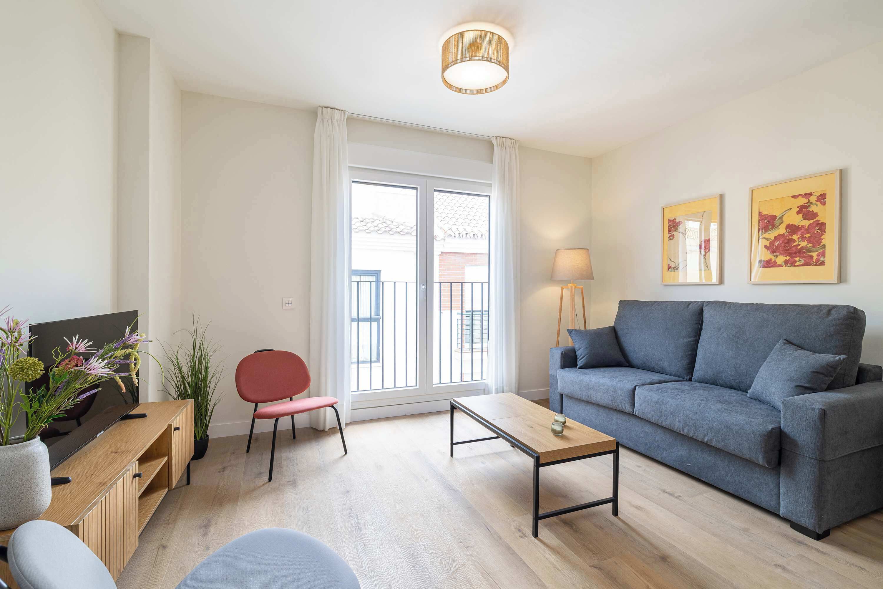 Ein modernes Wohnzimmer mit grauem Sofa, einem Holztisch, einem roten Sessel, einem Fernseher und einem großen Fenster mit Vorhängen.